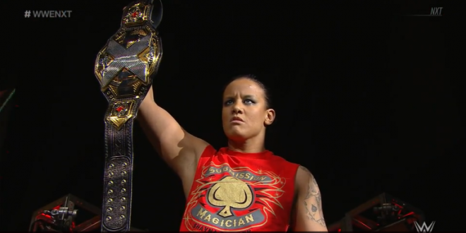 Shayna Baszler scowls, holding up her nXt Women's Championship, at the announce booth, on March 27th.