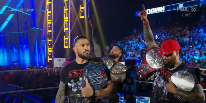 Roman Reigns wears his Universal Championship over his left shoulder, and his WWE Championship around his waist. He is wearing a promo t-shirt for his group, the Bloodline. He is standing in the middle of the wrestling ring with a microphone in his right hand. Behind him are his cousins, Jimmy and Jey Uso, also wearing their Tag Team Championship belts around both shoulders. Jimmy is raising his hand up, one finger raised.