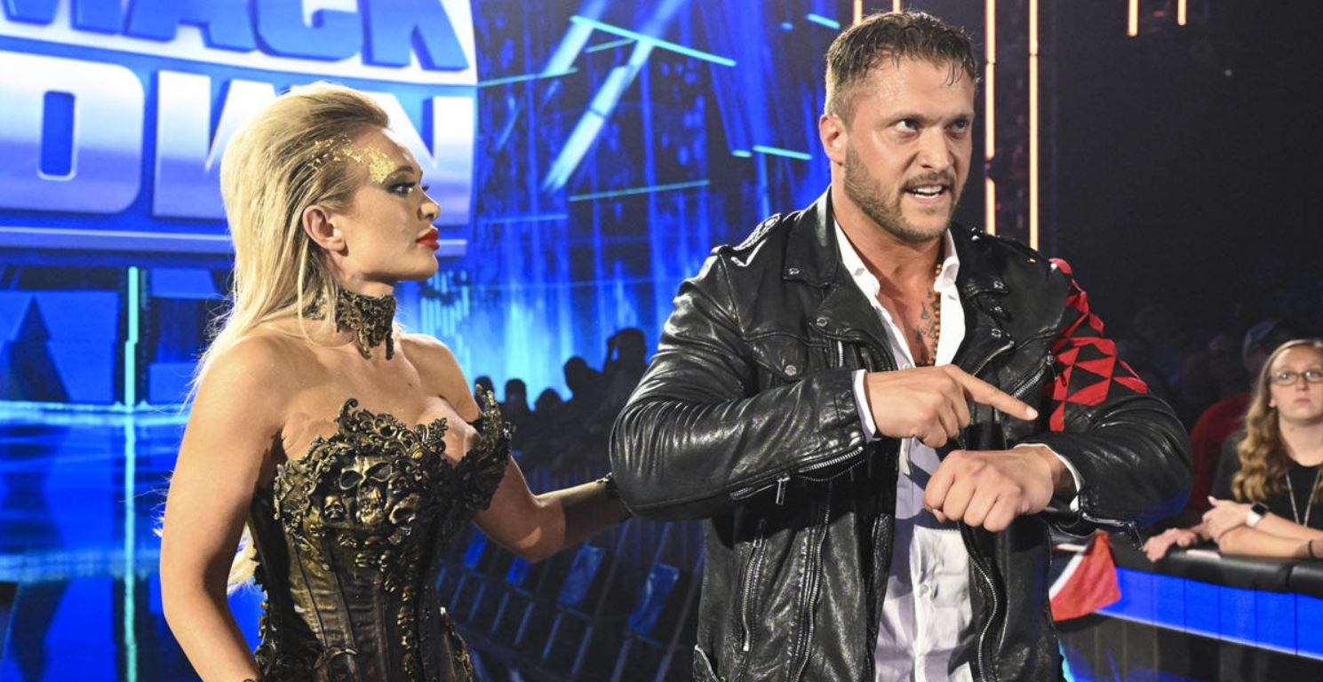 August 5, 2022, Friday Night Smackdown, Scarlett and Karrion Kross stand beside one another in the aisle, leading up to the wrestling ring. The blue and white Smackdown logo is visible behind them on the big screen. Scarlett has long, blonde hair, past her shoulders, dressed in a very fancy, shoulderless black and gold dress. Kross is wearing a black leather jacket with white collared shirt, pointing with his right hand to his left wrist, 