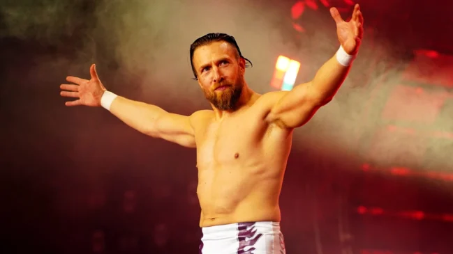 Bryan Danielson stands with his arms raised up in the air, palms facing out, greeting the crowd. He is not wearing a shirt. He has white pants. His brown hair is short, as is his beard.