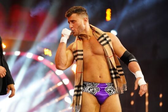 MJF stands on an entrance ramp during a wrestling show. He is looking to his right, wearing a gold and black scarf, and his purple wrestking trunks.
