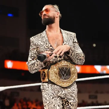 Seth Rollins stands in the wrestling ring at WWE Monday Night Raw, wearing the brand new World Heavyweight Championship around his waist. He is wearing a black and white patterned suit, with no shirt under his jacket. He has rose-tinted glasses on and his hair is up in a short ponytail.