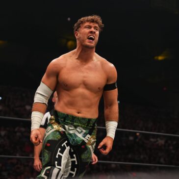 Will Ospreay stands looking out at the crowd. He is not wearing a shirt. He has white tape wrapped around his wrists. He is wearing dark green pants with a stylized pattern in white on them.