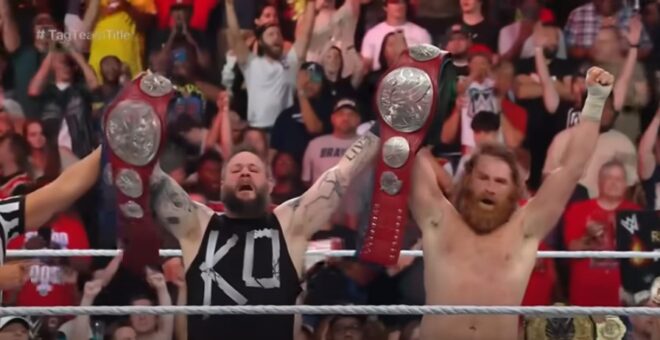 At left, Kevin Owens' right arm is raised in victory by the referee. Owens holds his WWE Raw Tag Team Championship in the same hand. He is wearing his trademark black KO cutoff t-shirt. Zayn is at right, holding his WWE Raw Tag Team Championship in his right hand, which is held by Owens. Both men are happy after winning a match over Judgment Day.